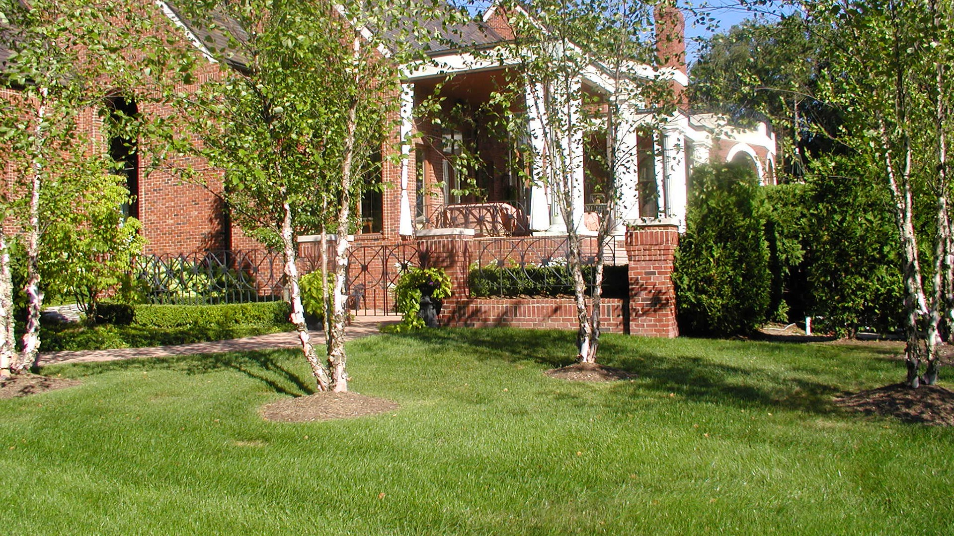 A red-brick building with white columns, surrounded by neatly trimmed grass and birch trees under a clear blue sky.