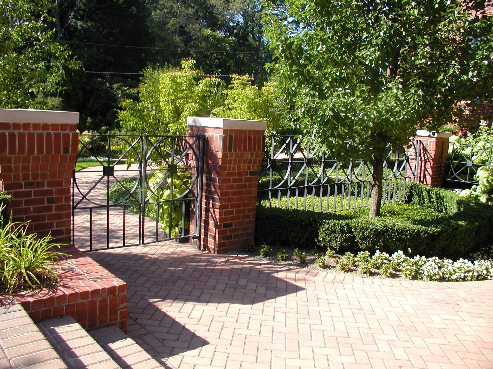 A brick pathway leads through an open wrought iron gate, surrounded by lush greenery and neatly trimmed hedges under a clear sky.