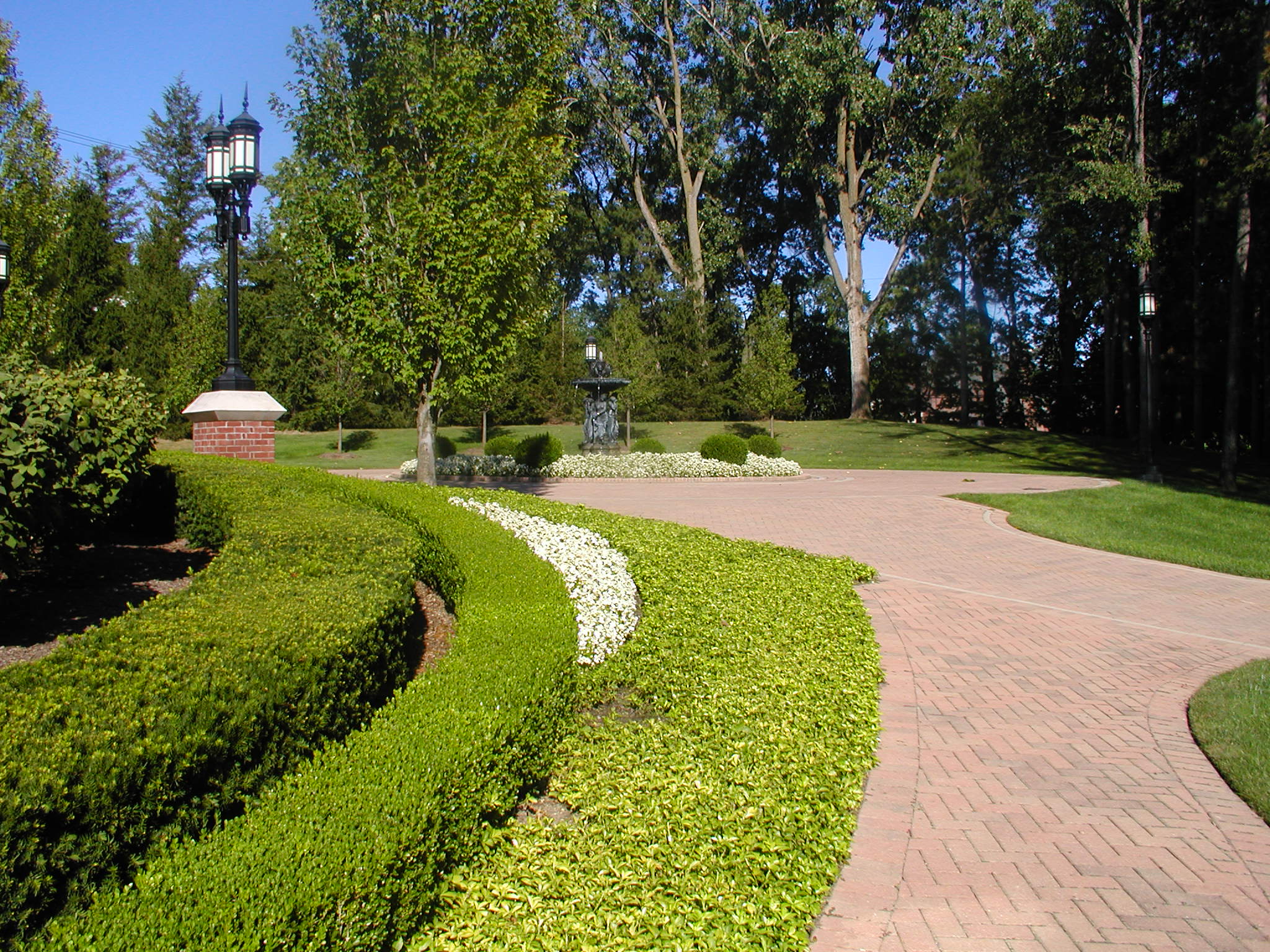 A landscaped garden features a curved brick walkway, manicured bushes, lampposts, and tall trees, creating a serene and inviting outdoor space.
