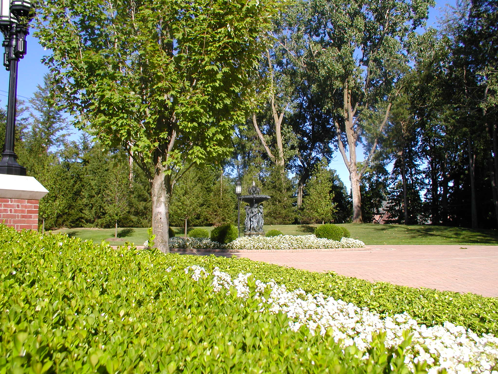 A peaceful garden with vibrant greenery, trees, a decorative fountain, and a brick path under a clear blue sky.
