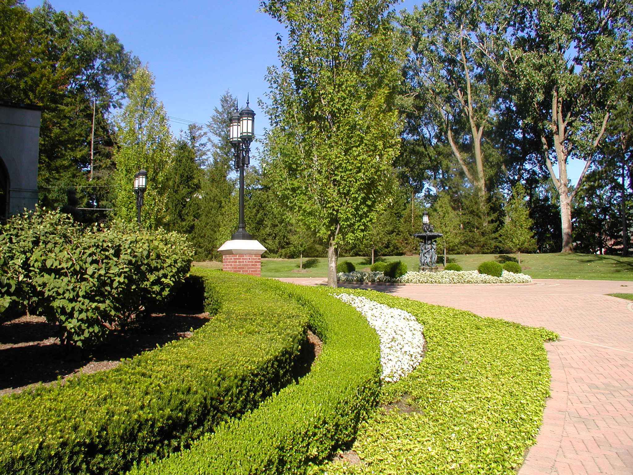A serene garden scene with trimmed hedges, colorful flowers, classic lamp posts, and a central fountain under a clear blue sky.