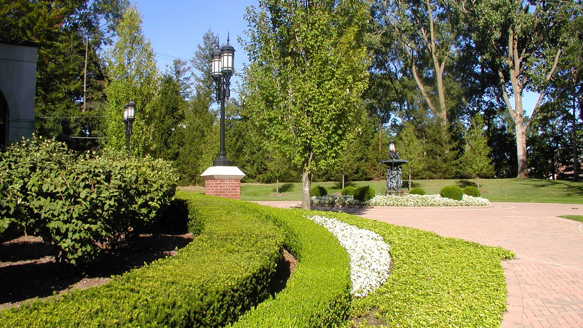The image shows a landscaped garden with a decorative fountain, brick path, neatly trimmed hedges, trees, and vintage-style street lamps.