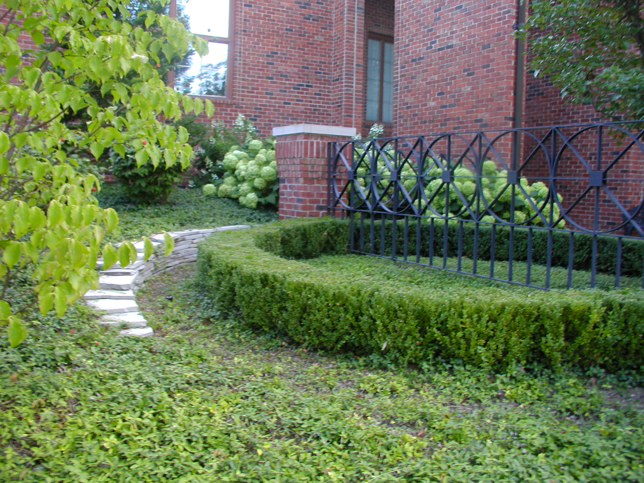 A neatly landscaped garden with brick walls, green shrubs, and a decorative metal fence alongside a stone walkway leads to a building entrance.