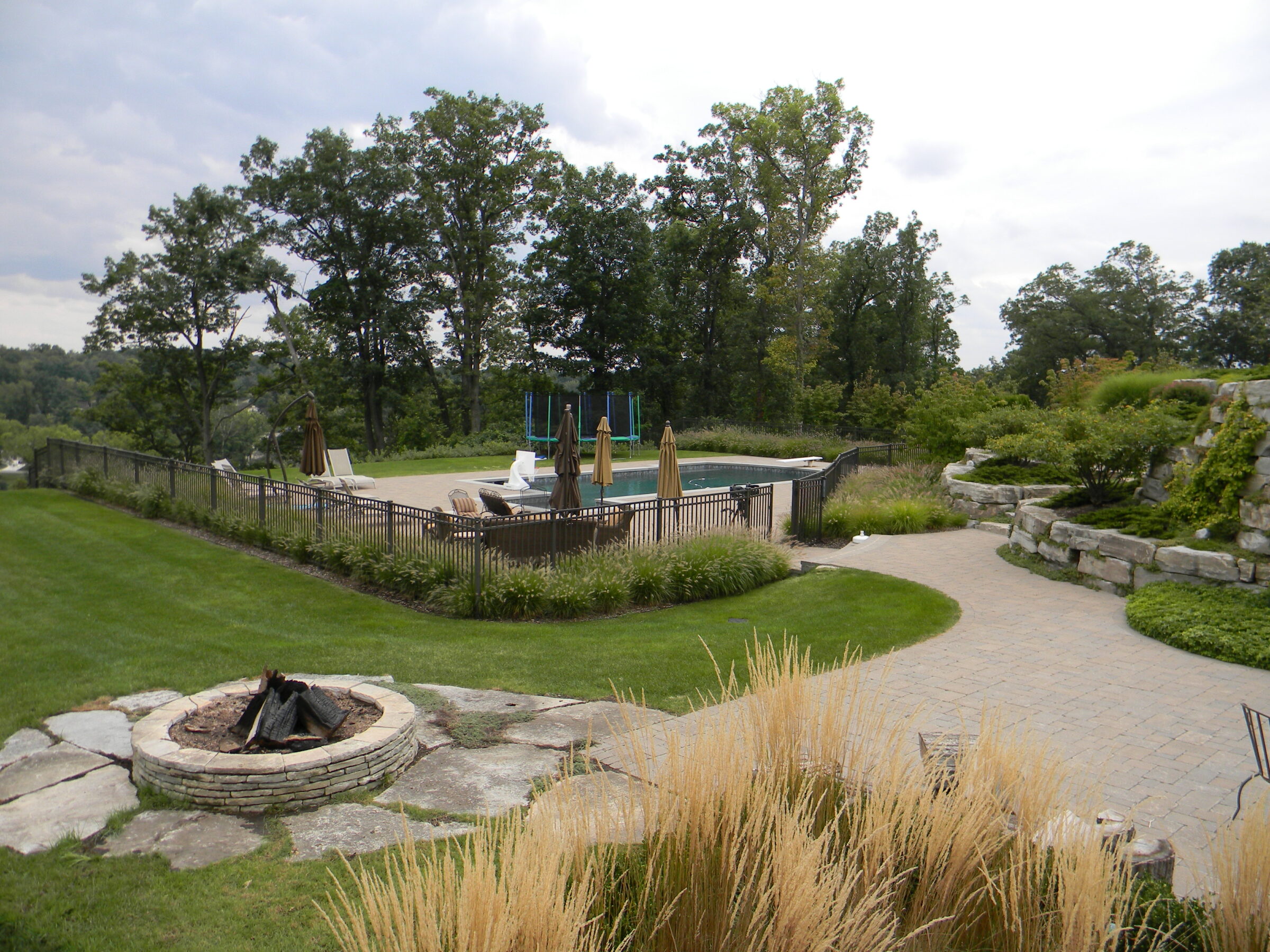 A serene backyard features a pool, fire pit, and stone path, surrounded by greenery and trees under a cloudy sky.