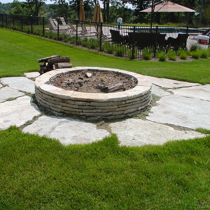 A stone fire pit with logs is set on paved stones, near a fenced pool area with chairs and umbrellas.