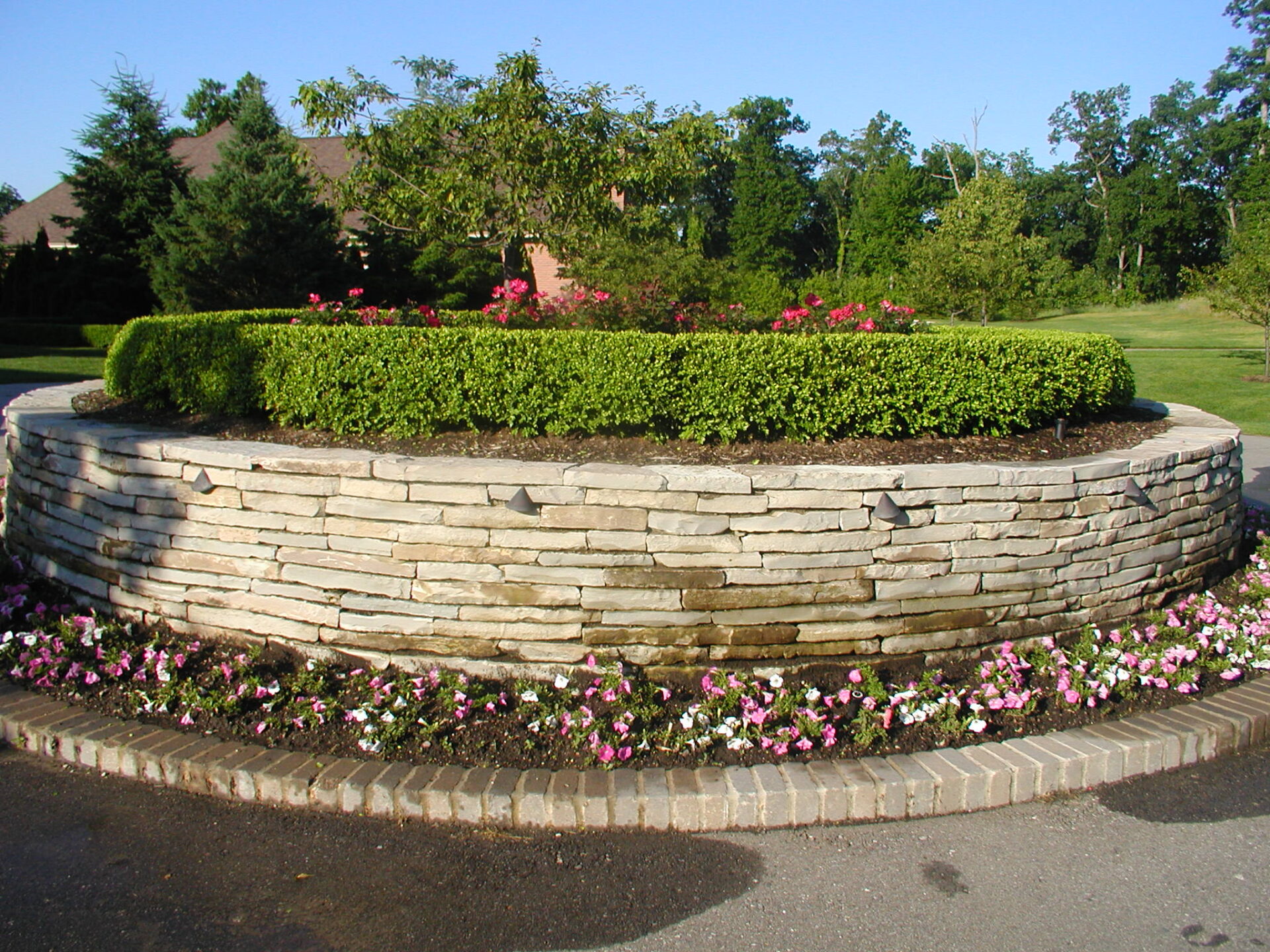 A circular stone planter features neatly trimmed greenery and vibrant flowers, surrounded by a brick border. Trees and shrubs fill the background.
