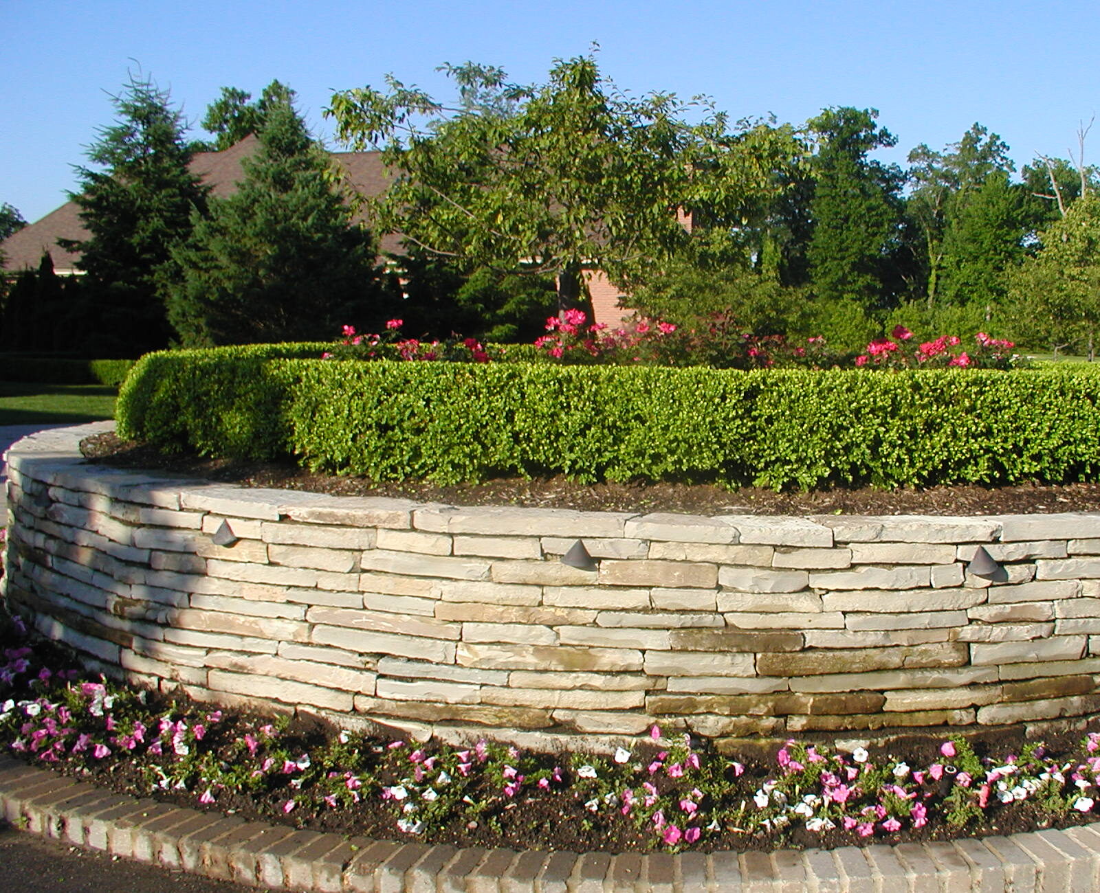 A circular stone planter features neatly trimmed greenery and vibrant flowers, surrounded by a brick border. Trees and shrubs fill the background.