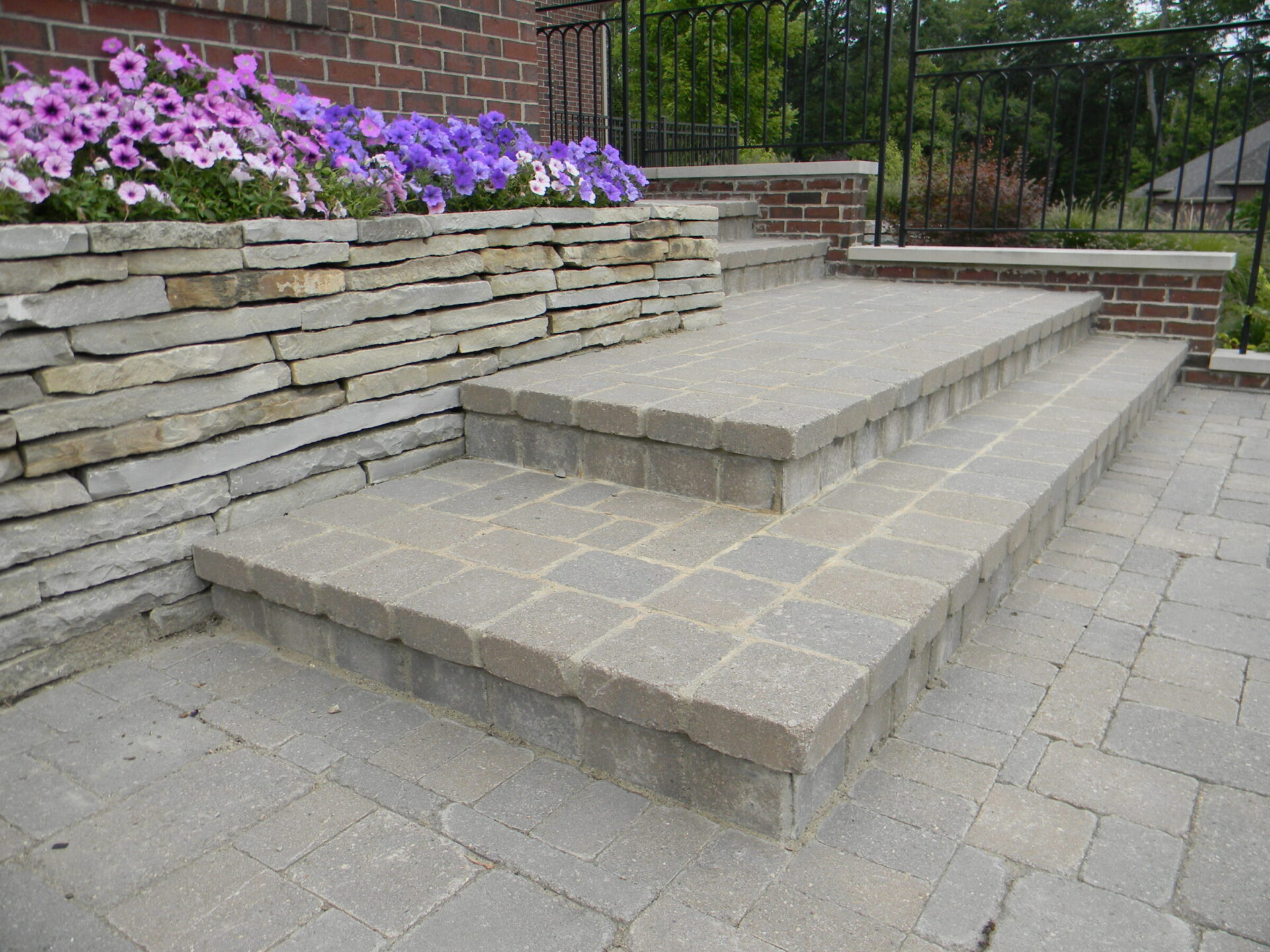 Brick and stone steps with colorful flowers border a garden path. The scene is peaceful, framed by greenery and a metal railing.