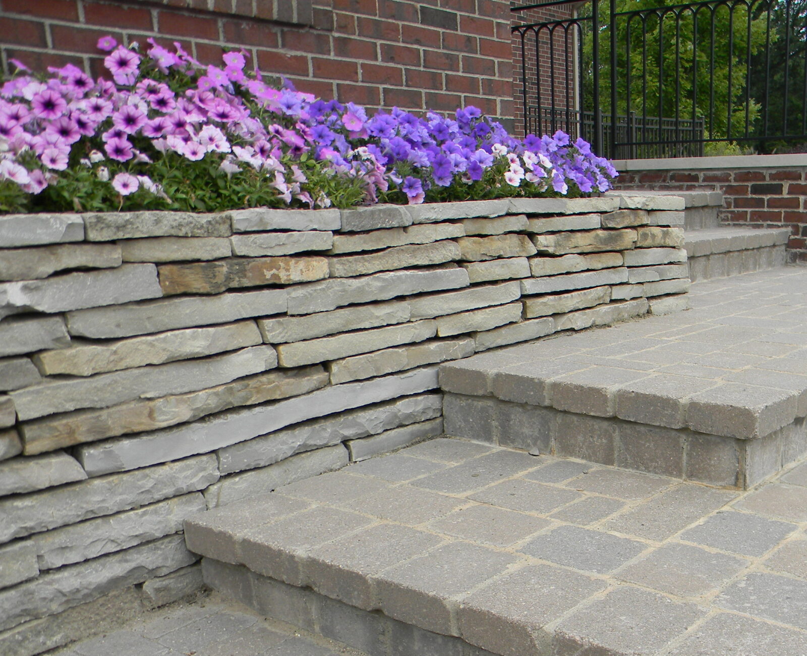 Brick and stone steps with colorful flowers border a garden path. The scene is peaceful, framed by greenery and a metal railing.