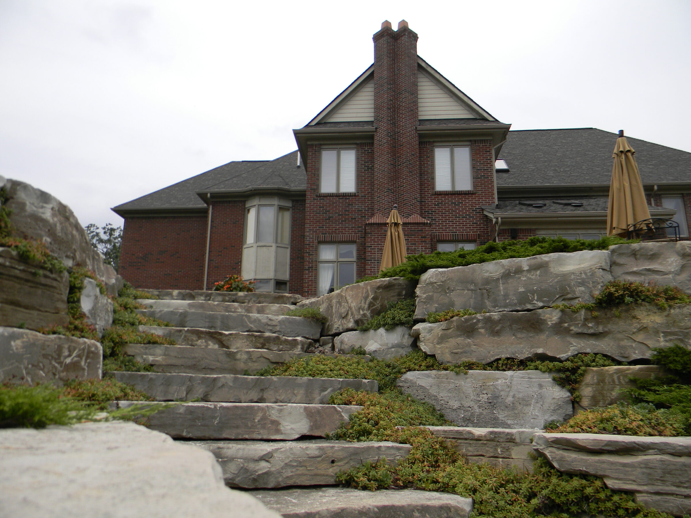 A brick house with multiple chimneys, stone steps, greenery, and outdoor umbrellas in the yard, under a cloudy sky.