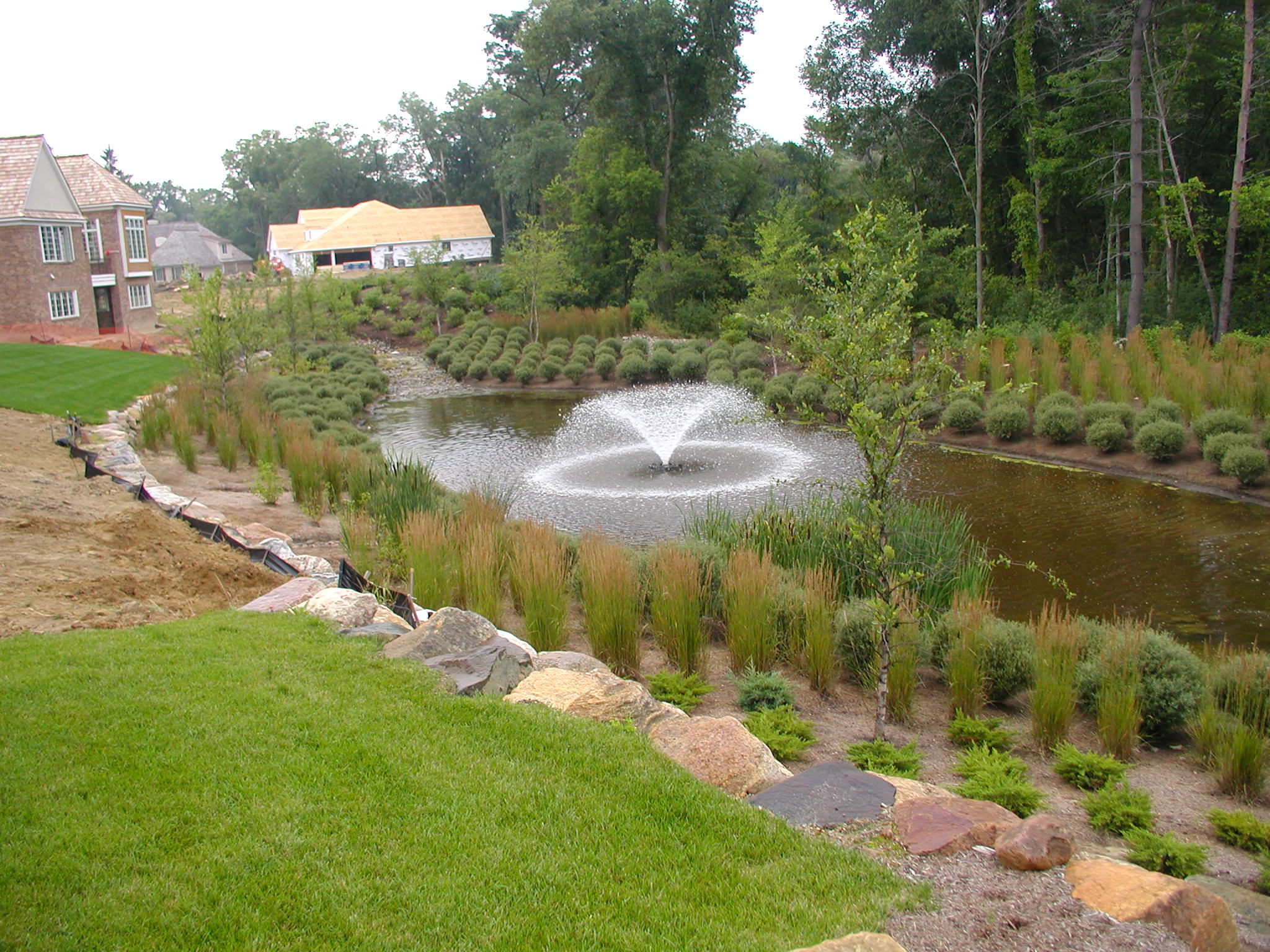 A landscaped garden features a pond with a central fountain. Nearby, there are houses under construction amid lush greenery and trees.