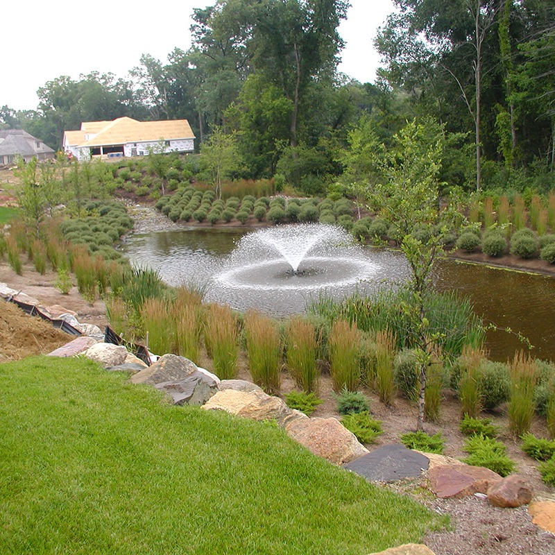 A serene garden features a small pond with a central fountain, surrounded by lush greenery and a distant house under construction.