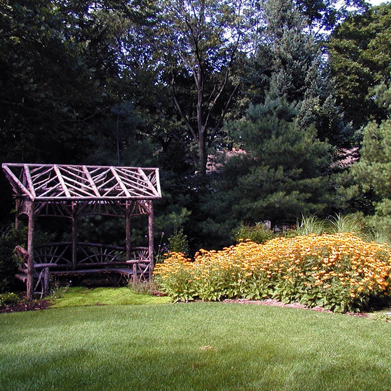 A rustic wooden bench sits in a lush garden with vibrant orange flowers and dense trees under a clear sky.