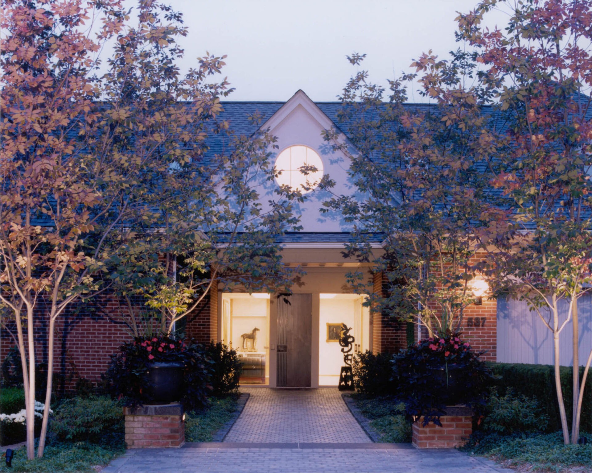 A house entrance is framed by trees and greenery. Warm light spills from the doorway, highlighting modern sculptures inside.