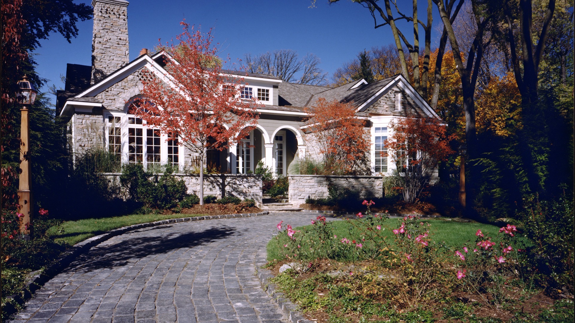Charming stone house with arched entry, surrounded by autumn trees and a cobblestone path, under a clear blue sky.