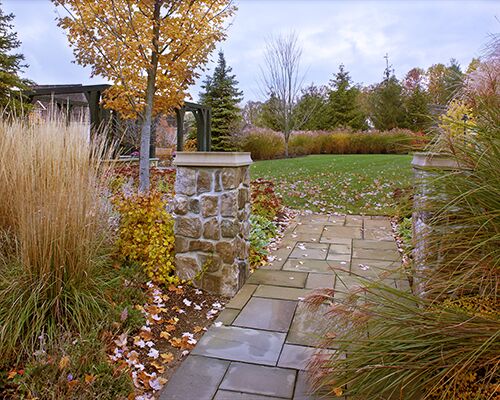 A stone path leads through an autumn garden with trees and shrubs, featuring a pergola and pillars under a cloudy sky.