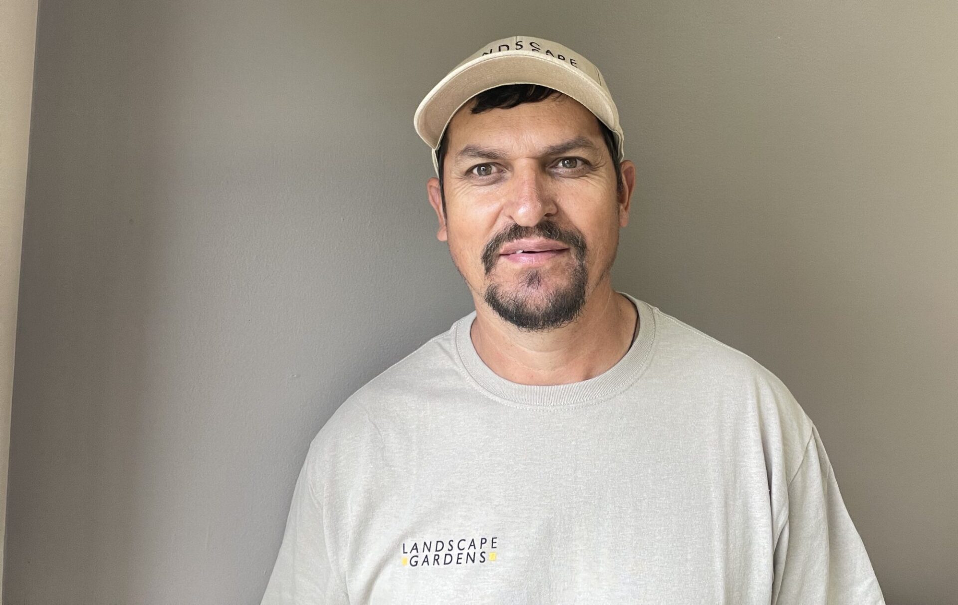 A person wearing a "Landscape Gardens" shirt and cap stands indoors against a plain wall, with a small landscape painting on the side.