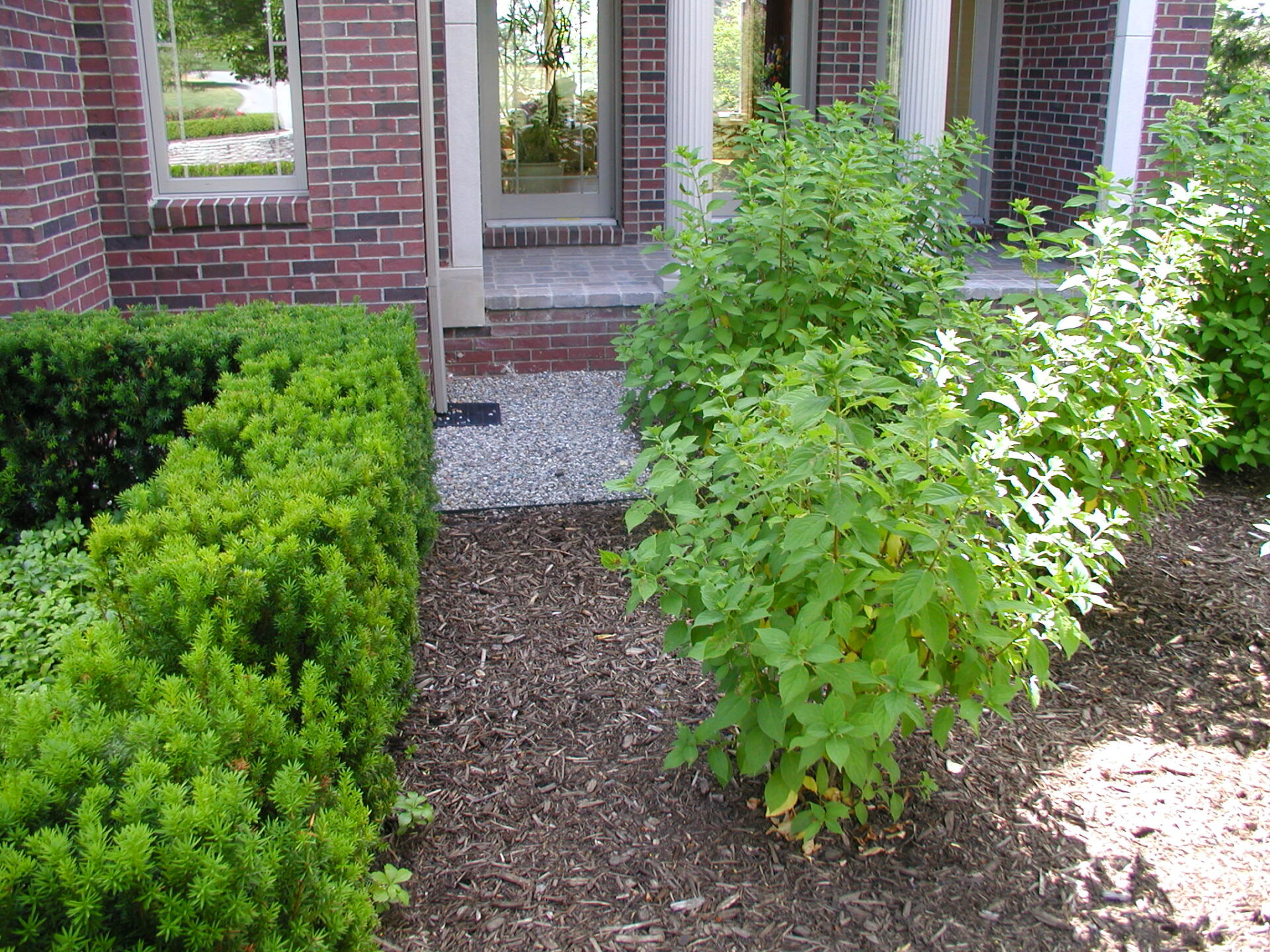 Red brick building features lush greenery and neatly trimmed shrubs, with a gravel path leading to a doorway with white columns.