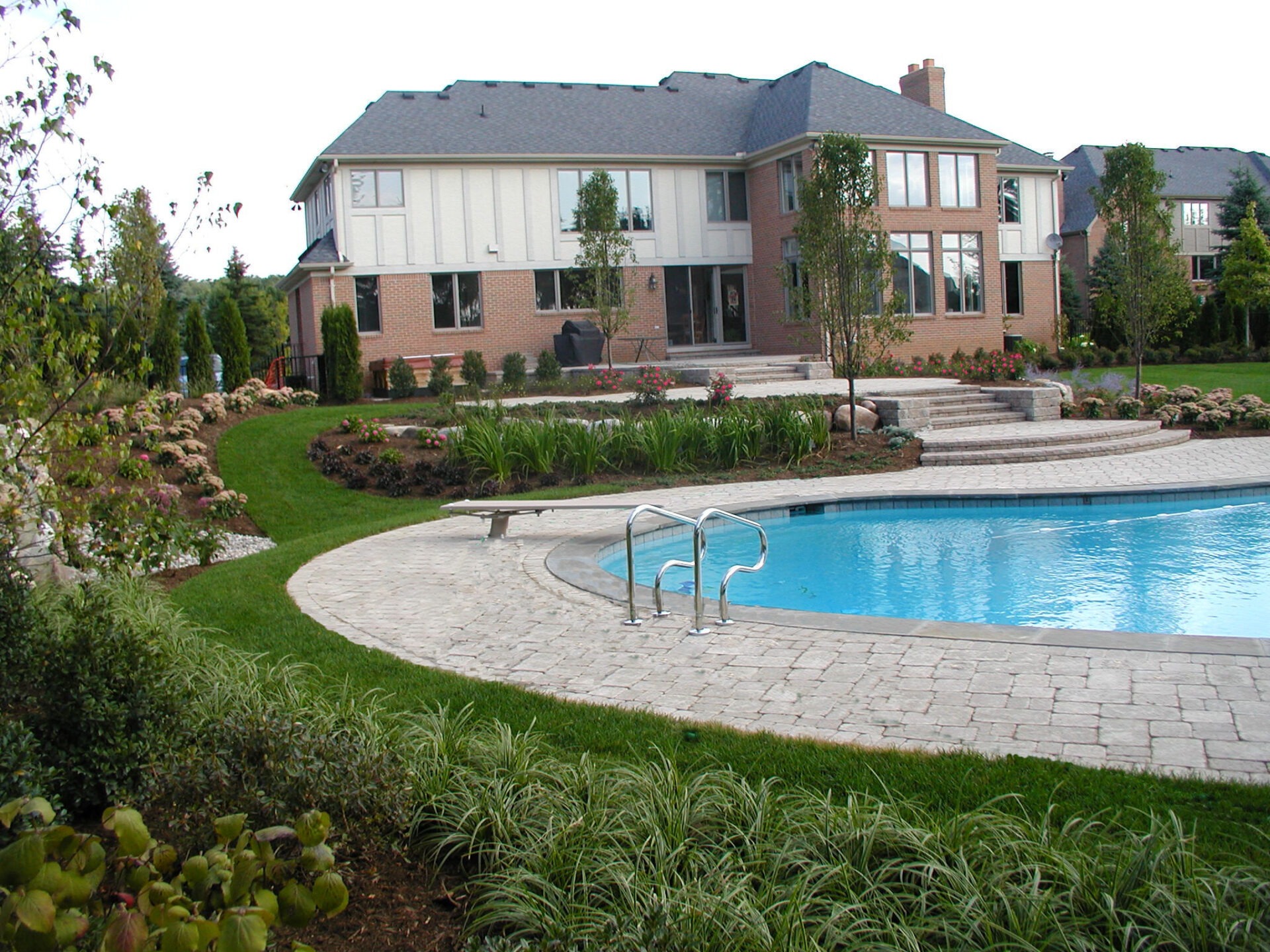 A brick house with a large swimming pool, surrounded by lush green landscaping and stone pathways, under a clear blue sky.