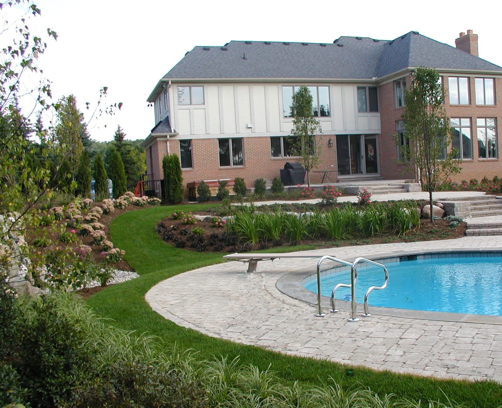 A brick house with a large swimming pool, surrounded by lush green landscaping and stone pathways, under a clear blue sky.
