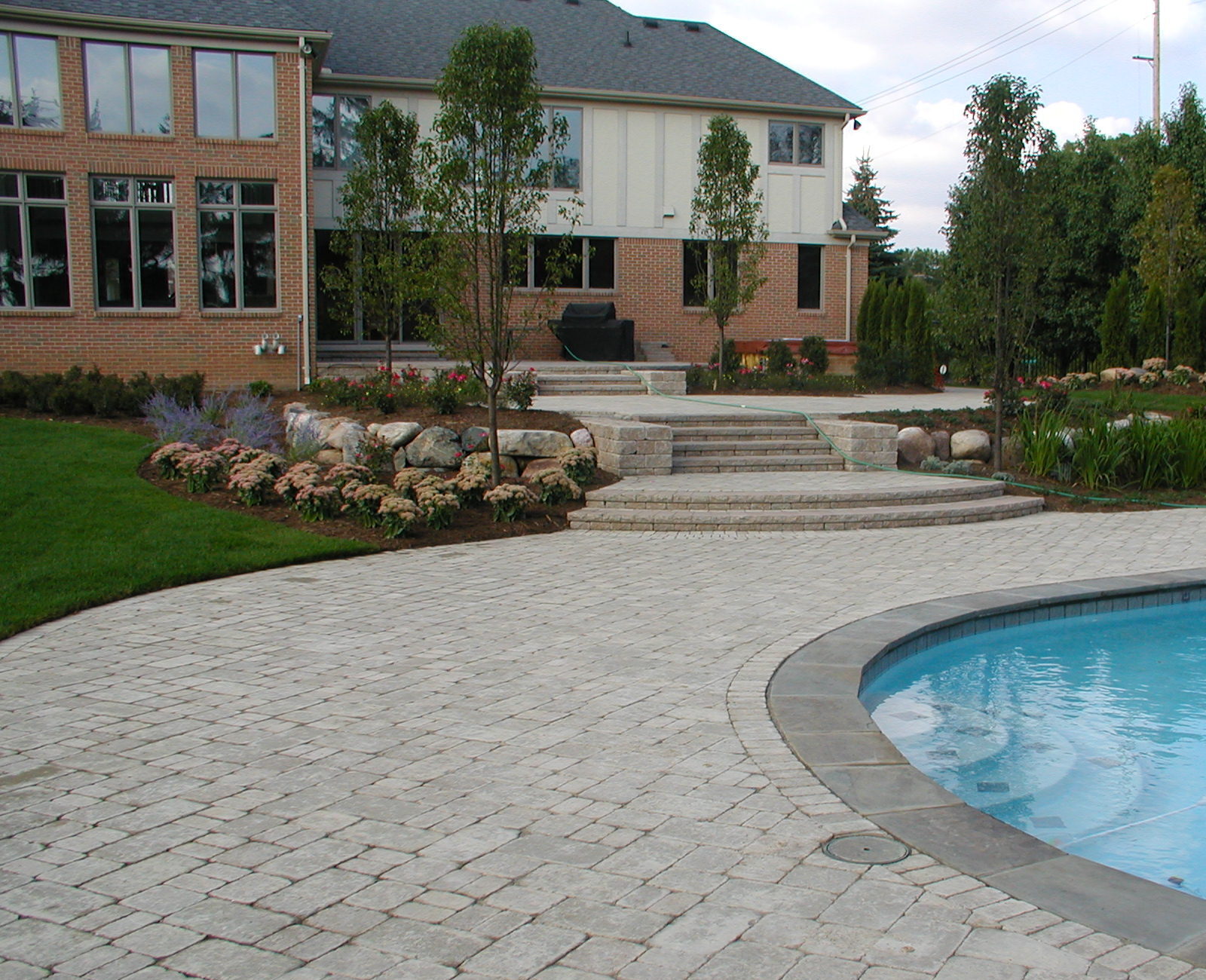 Spacious backyard with a curvy pool, well-maintained garden, brick house, and paved patio, surrounded by trees and neatly arranged flowers.