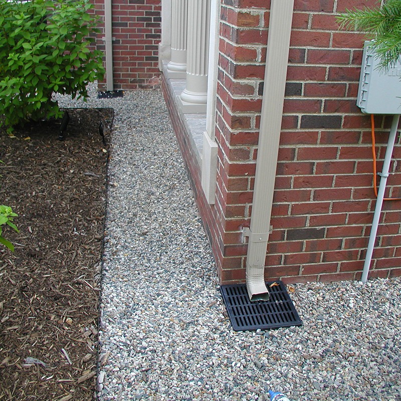 Brick building corner with a downspout draining into a gravel area. Bushes and a mulch bed line the path beside it.