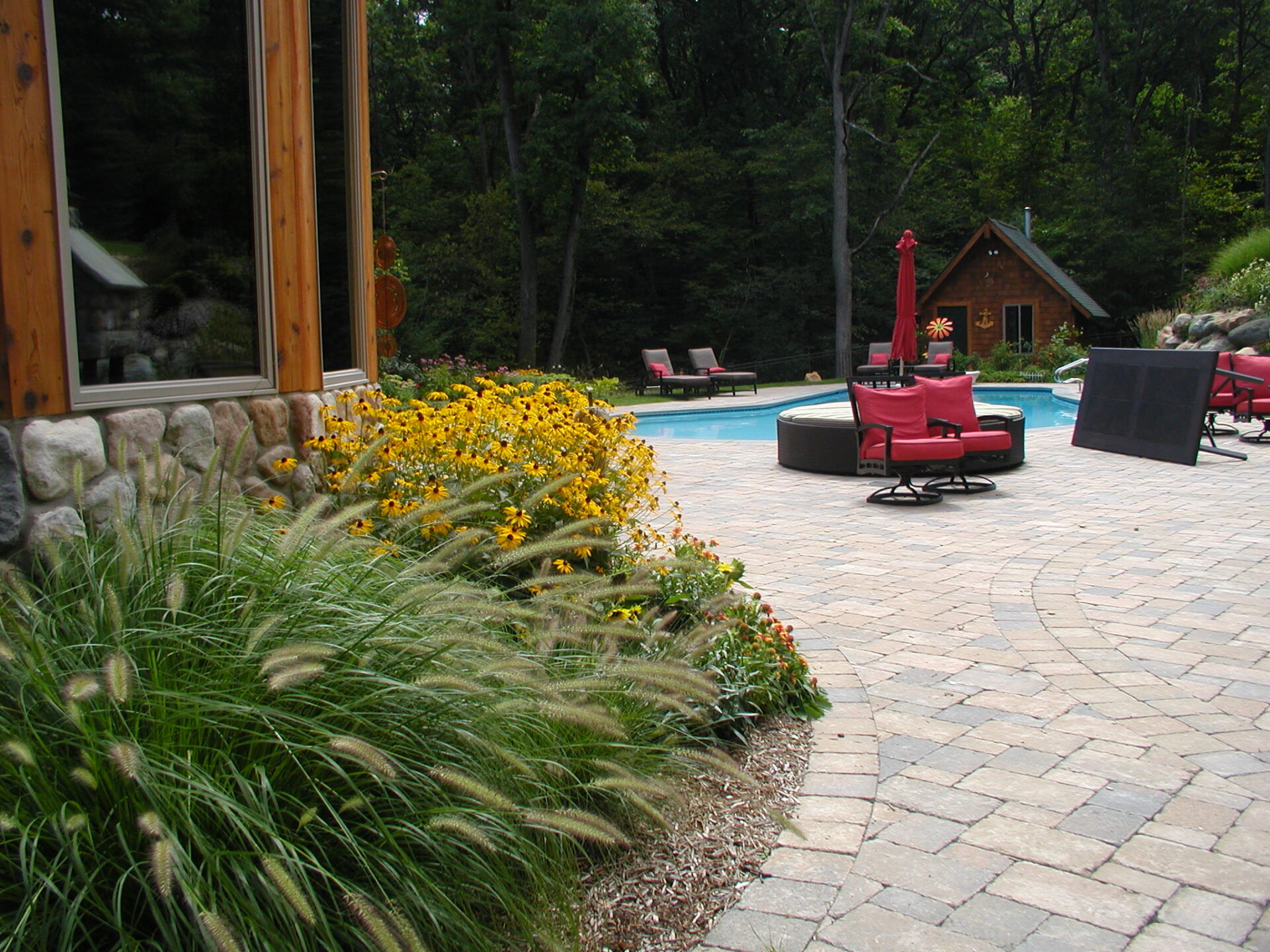 Garden with vibrant flowers and lush greenery surrounds a pool area with red-cushioned seating and a quaint wooden shed in the background.