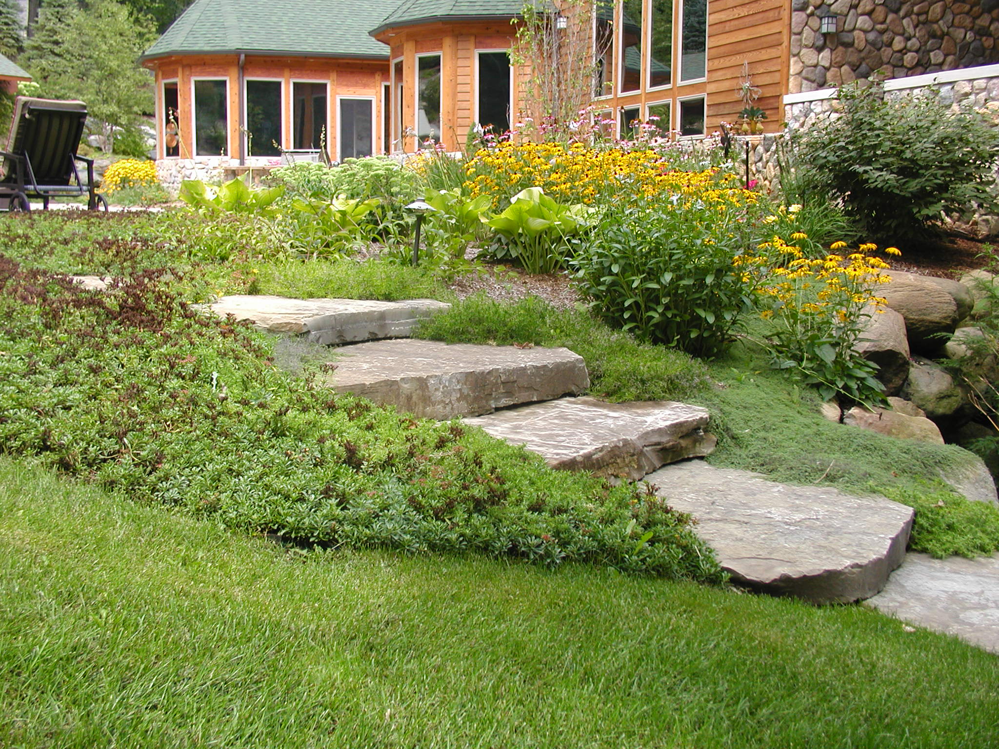 Wooden house with stone accents, surrounded by lush garden featuring yellow flowers. Stone path leads through greenery. No recognizable landmarks or people present.