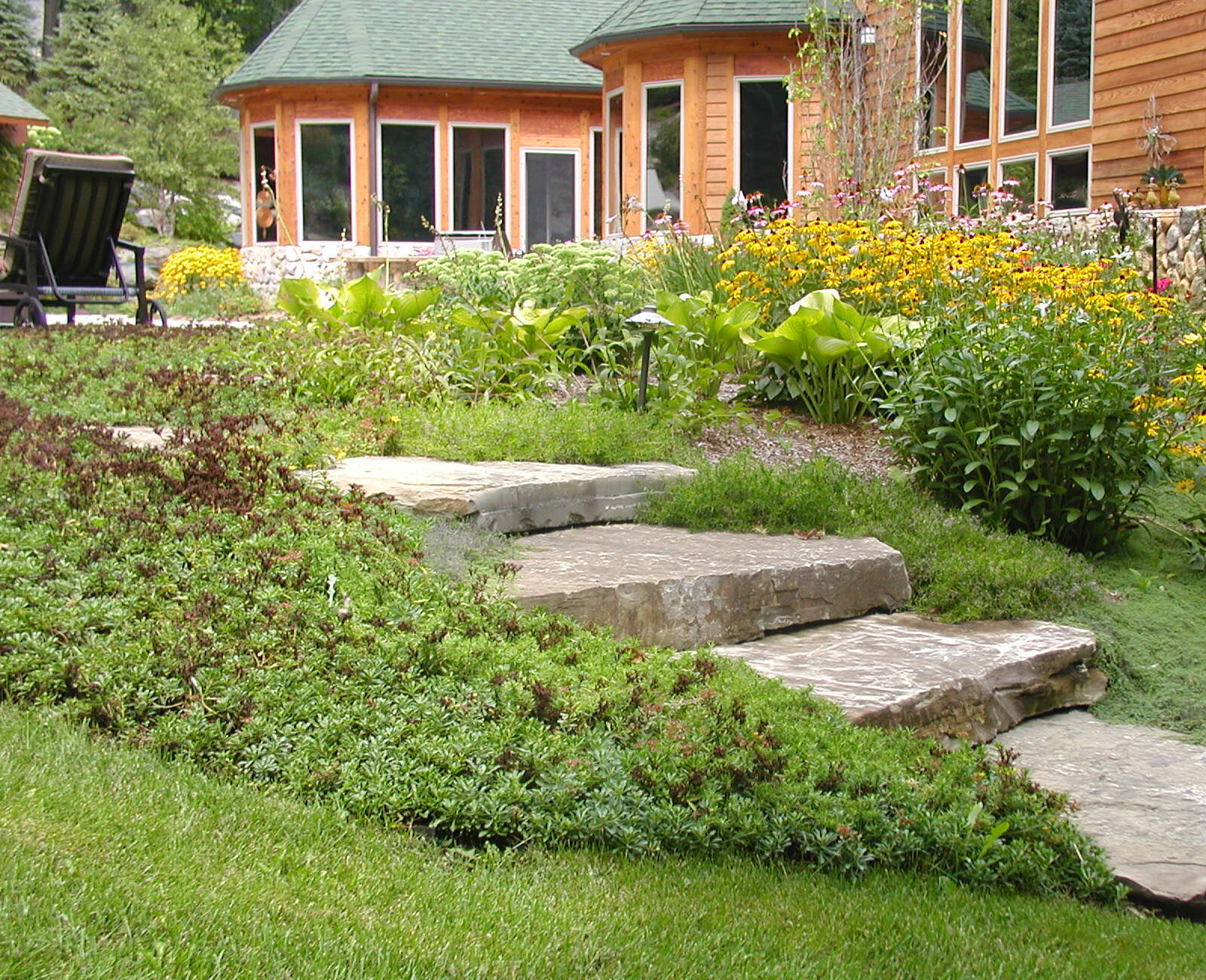 A landscaped garden features stone steps, lush greenery, and vibrant flowers leading to a wooden house with large windows and a green roof.