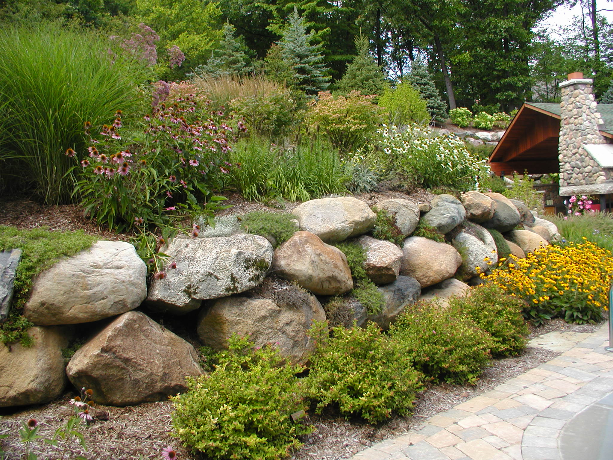 A beautifully landscaped garden features vibrant flowers, lush greenery, and a stone retaining wall beside a rustic wooden pavilion in a serene setting.
