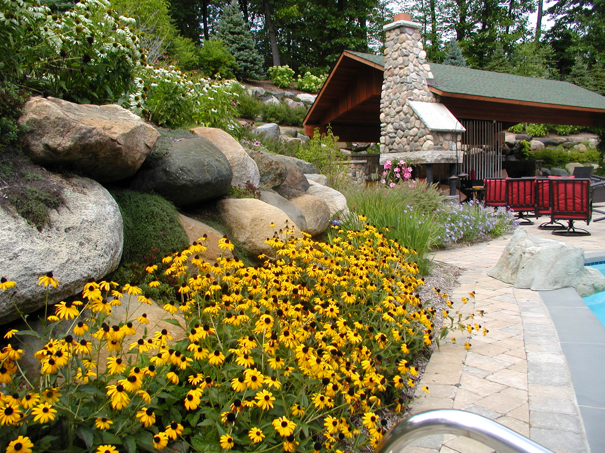 Colorful garden with vibrant flowers, stone pathway, rustic pavilion with stone fireplace, surrounded by lush greenery, chairs, and a swimming pool nearby.
