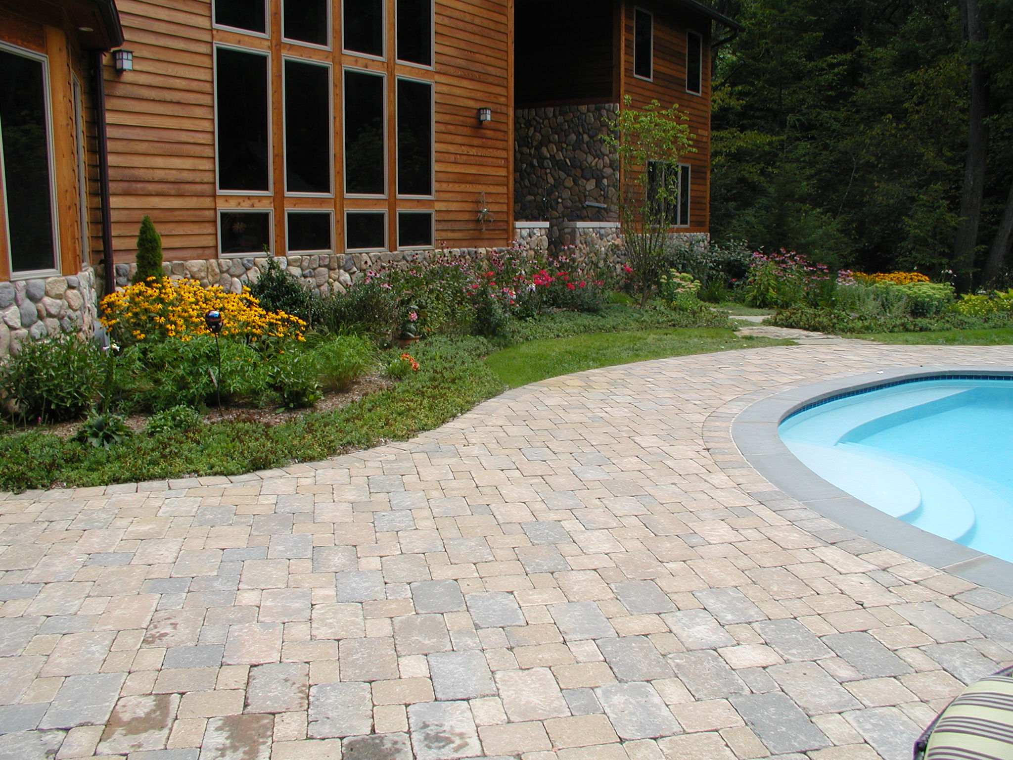 Wooden house with stone details, adjacent to vibrant garden, paved patio, and curved swimming pool. Surrounded by lush trees, serene setting.