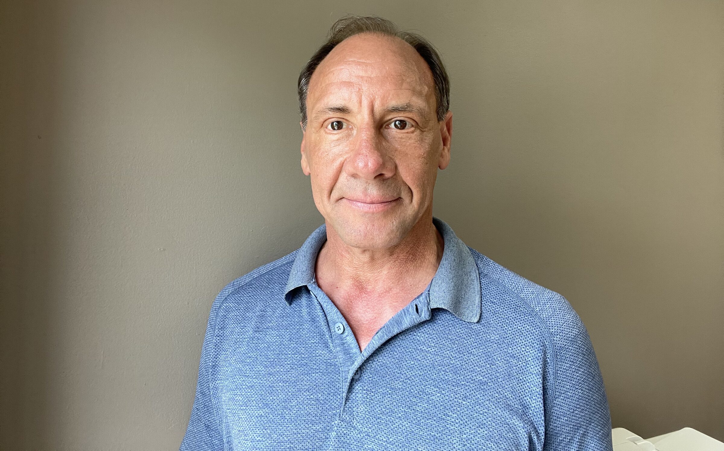 A person in a blue collared shirt stands against a plain, neutral-colored wall, looking directly at the camera with a subtle smile.