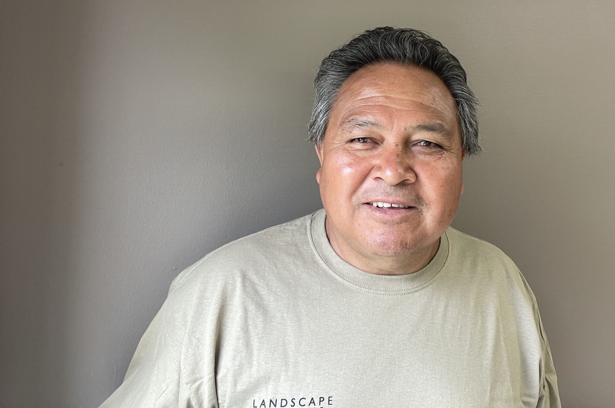 A person wearing a beige shirt smiles against a neutral, light brown wall. The shirt has partial visible text reading "LANDSCAPE".