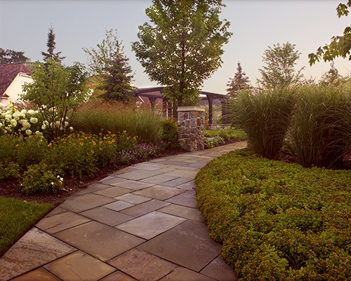 A stone path winds through a lush garden with trees, flowers, and a small stone pillar under a clear sky.