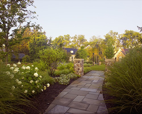 A stone pathway leads through a lush garden with flowering bushes and trees, bordered by stone pillars, under a clear blue sky.