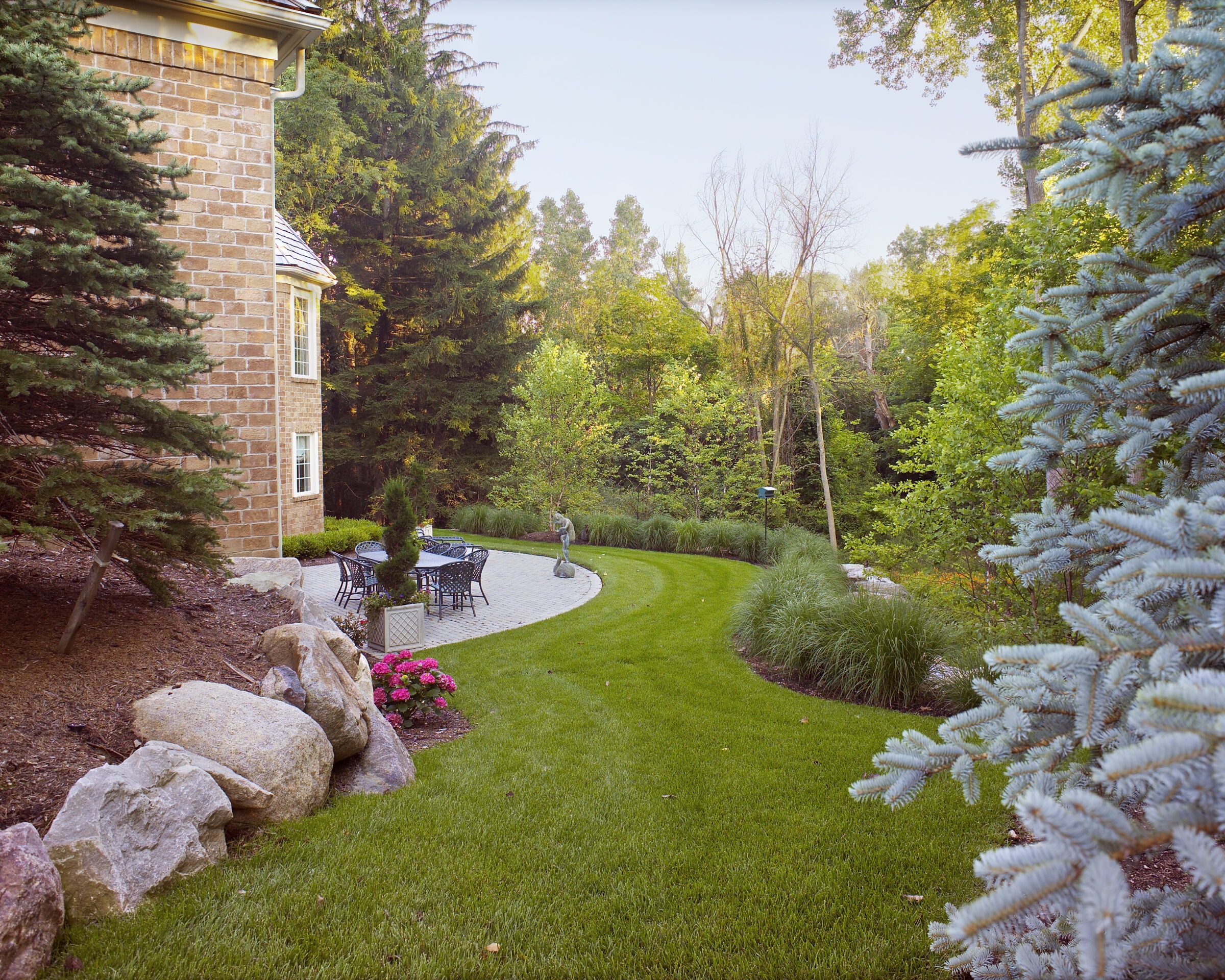 Brick house beside well-manicured lawn and patio. Outdoor furniture and statue amid lush green trees and landscaped garden, creating a serene atmosphere.