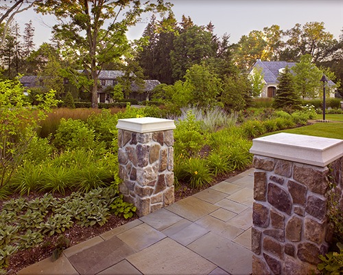 Stone pillars flank a landscaped garden path, surrounded by lush greenery and trees, leading to a house in the background under a clear sky.