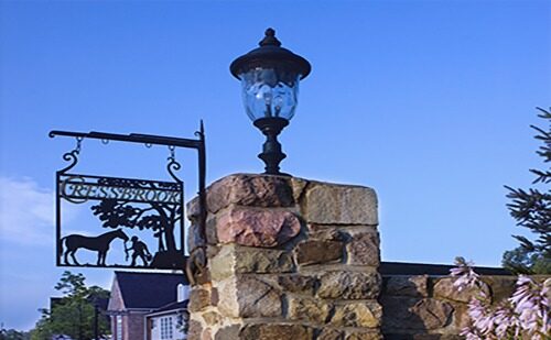 Stone pillar with an ornate lantern and horse-themed sign reading "Cressbrook." Clear blue sky and partial view of surrounding greenery.