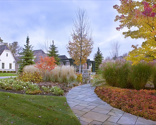 A stone path winds through a landscaped garden with colorful autumn foliage, leading towards a pavilion. A house is visible in the background.