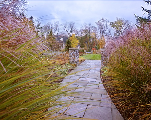 A stone pathway winds through a landscaped garden with tall grasses, leading towards a small building amid autumn trees and overcast skies.