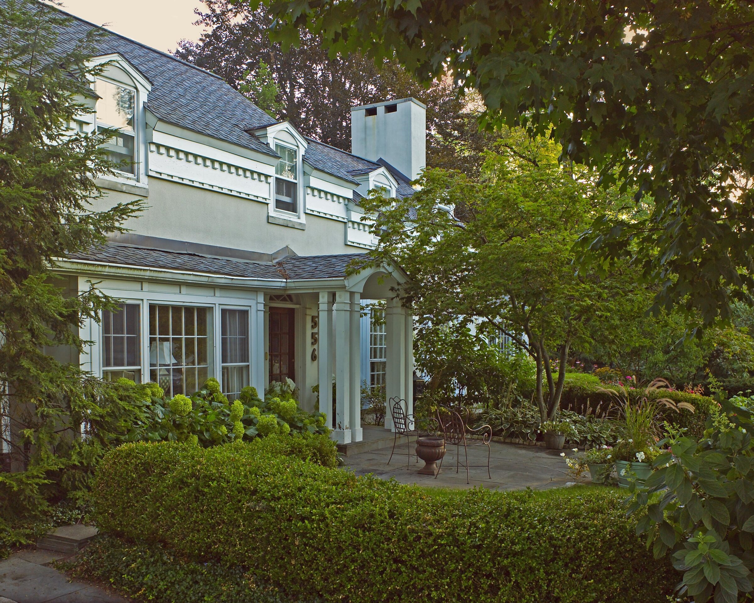 Charming two-story house with dormer windows, surrounded by lush greenery and garden. Wrought iron chairs sit on a stone patio, creating a cozy atmosphere.