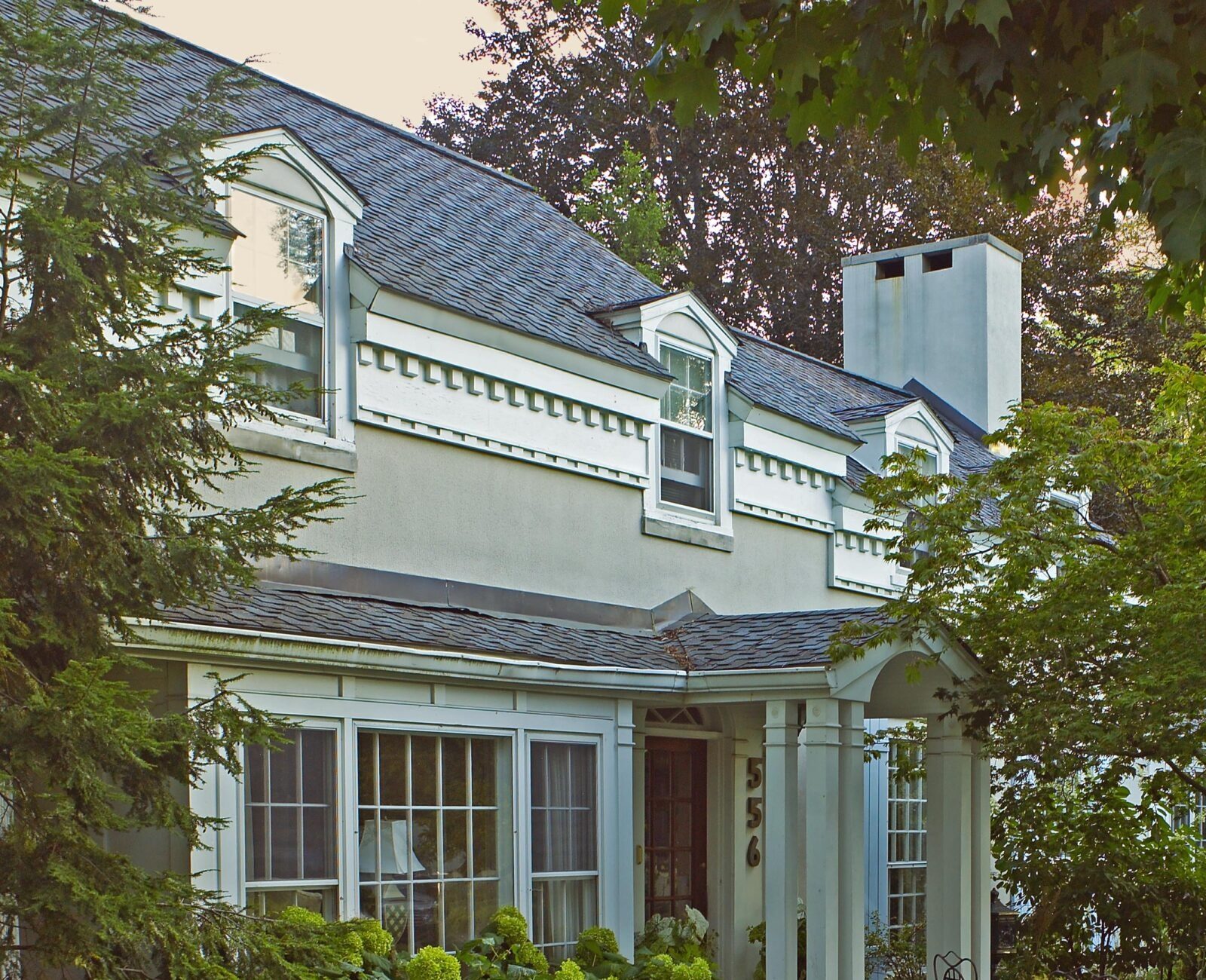 Charming two-story house with dormer windows, surrounded by lush greenery and a cozy patio, creating a serene and inviting garden atmosphere.