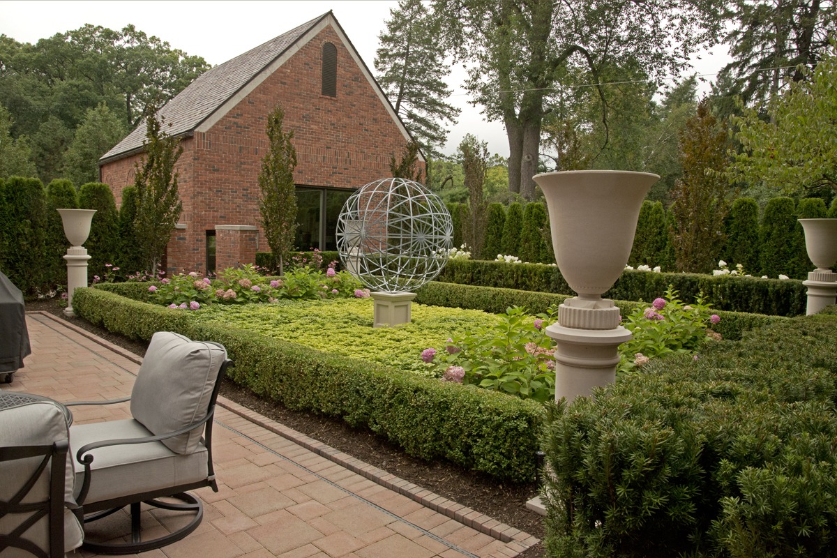 A beautifully manicured garden with a brick building, metal sphere sculpture, potted plants, and comfortable patio furniture.