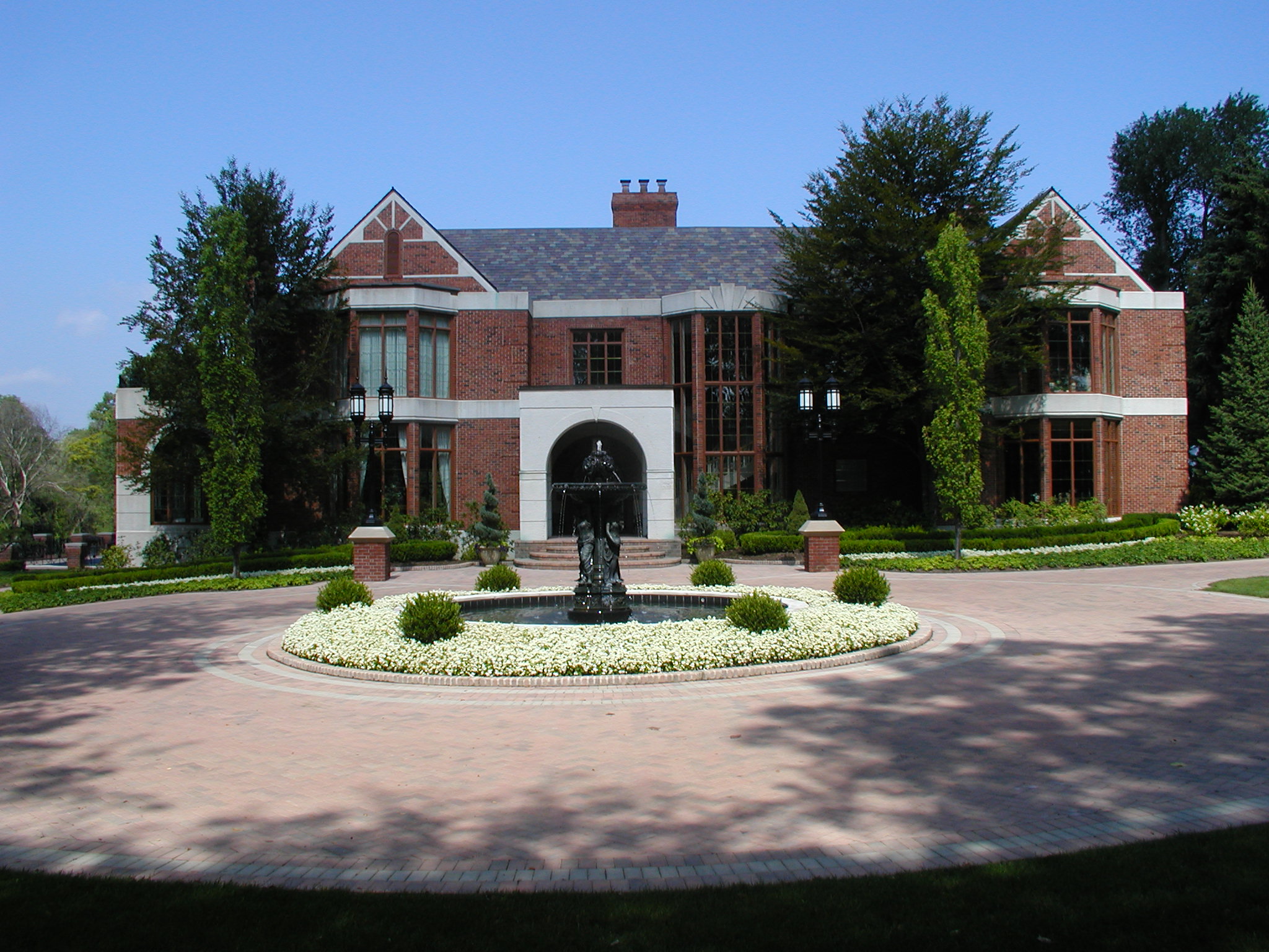 Grand red-brick mansion with circular driveway, central fountain. Surrounded by lush greenery and trees against a clear blue sky.