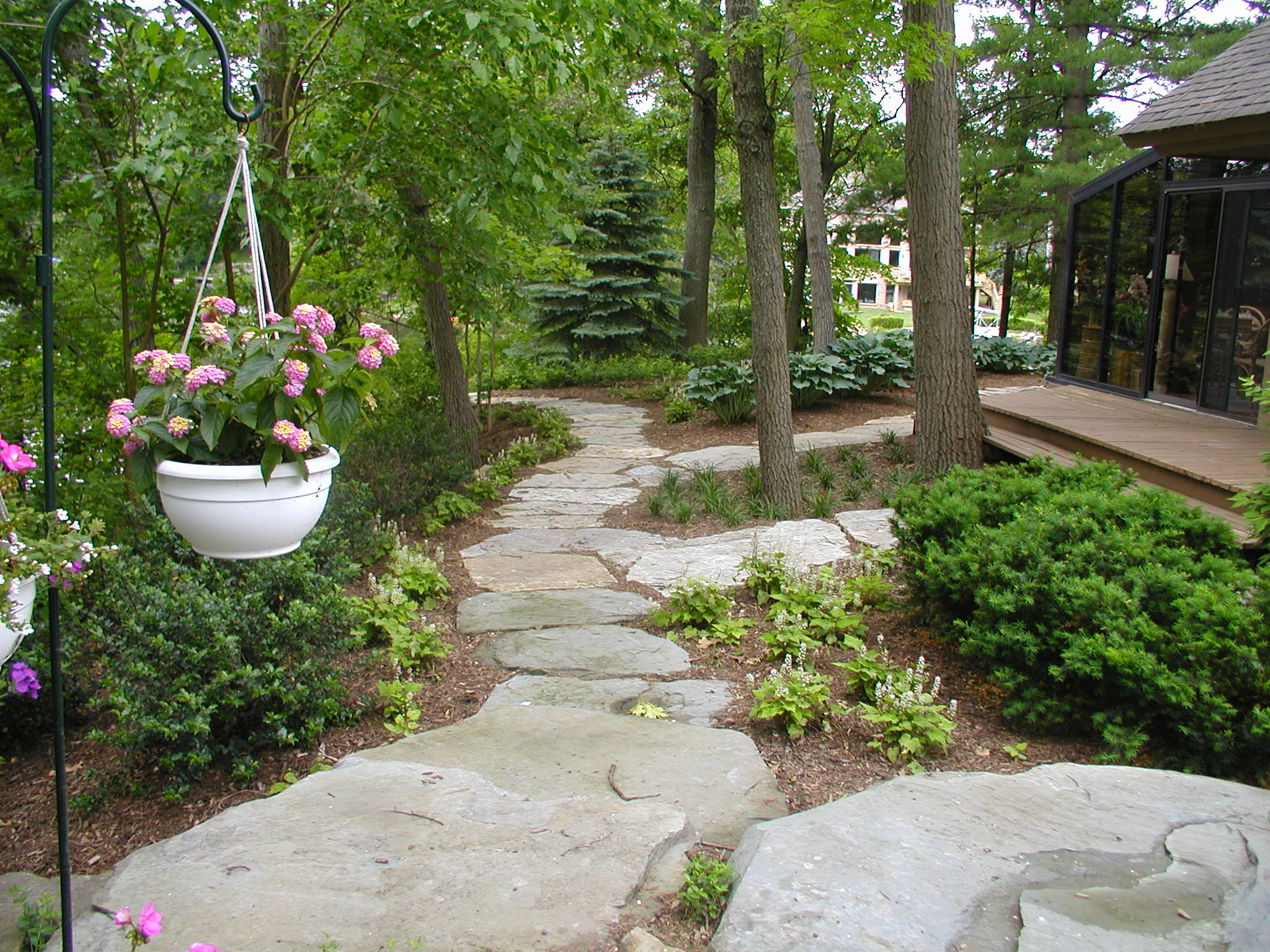 A serene garden with stone path, lush greenery, hanging flowers, and a wooden deck amidst tall trees. Calm, natural ambiance prevails.