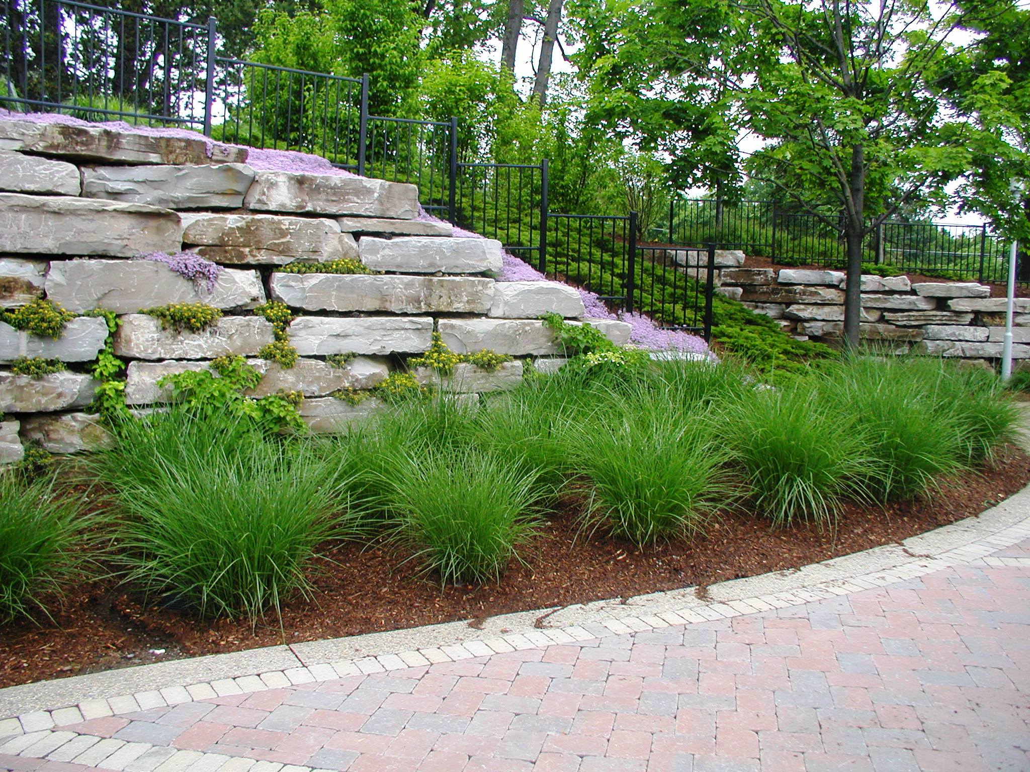 A stone retaining wall with lush greenery and flowering plants, bordered by a curved brick pathway in a landscaped garden.