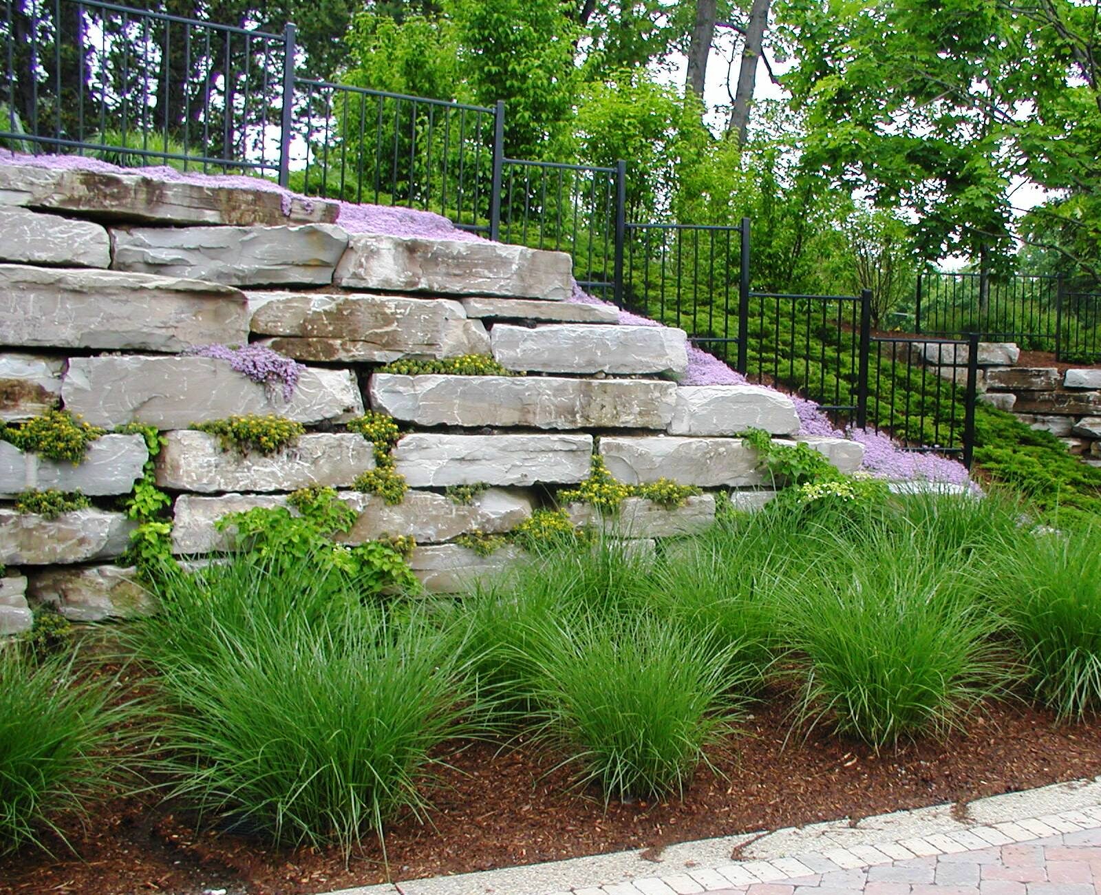 A landscaped garden with large stone retaining walls, purple flowers, ornamental grasses, and trees. Brick pathway curves through the lush greenery.