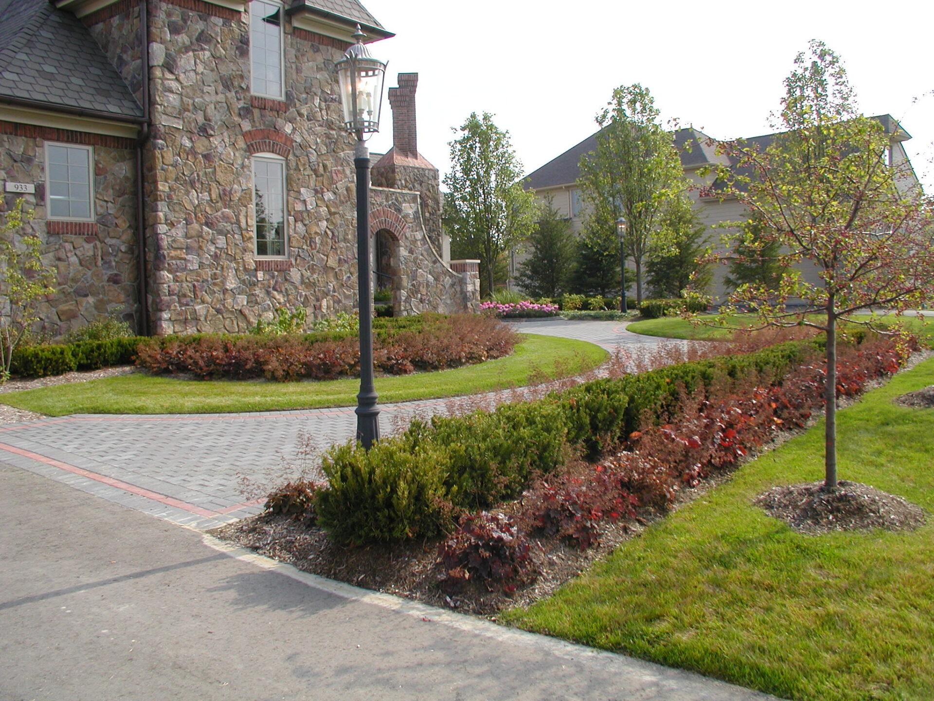 Stone house with a lamp post and manicured garden. Curved path with bushes and trees. Clear, sunny day.