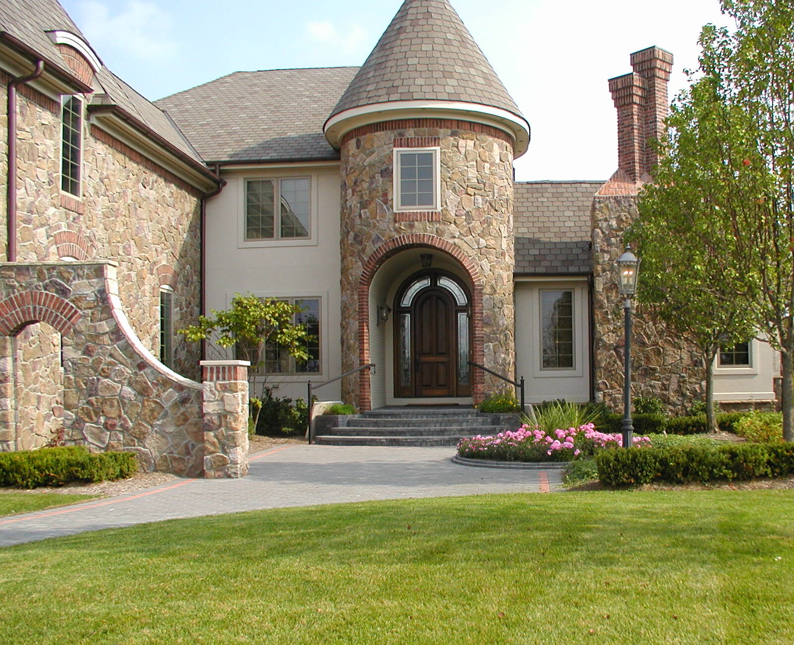 Stone house with turret, surrounded by lush green lawn and trees. Elegant entrance and well-maintained garden under clear blue sky.